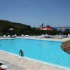 Piscina all'aperto con ombrelloni di paglia e lettini. Vista collinare e mare sullo sfondo.