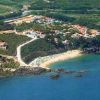 Vista aerea di un litorale con vegetazione, strutture turistiche, una piccola spiaggia e mare blu.