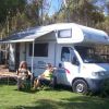 Due persone sedute davanti a un camper con tende aperte in un'area verde con alberi.