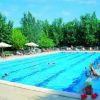Una piscina con diverse persone che nuotano e giocano, circondata da alberi e lettini.