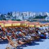 Spiaggia sabbiosa con file di ombrelloni gialli e sdraio. Sullo sfondo, una città sulla scogliera.