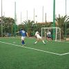 Partita di calcio su campo verde recintato, con due giocatori vicino alla porta e un portiere.
