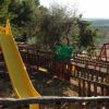 Un parco giochi recintato con uno scivolo giallo e altalene tra gli alberi.