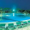 Grande piscina notturna con lettini, circondata da palme e capanne di paglia.