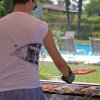 Una persona in maglietta e pantaloncini cucina alla griglia vicino a una piscina.