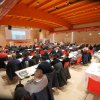 Conferenza con persone sedute a tavoli, schermo proiettato.