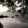 Un molo sull'acqua con persone sedute sotto un albero. In lontananza, una barca a vela sul lago.