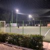 Un campo di calcetto illuminato di sera, con giocatori in azione e una recinzione verde attorno.
