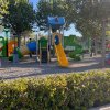 Un parco giochi con scivoli gialli e verdi, un'area in ghiaia e alberi intorno.