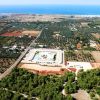 Vista aerea di una struttura circondata da campi e uliveti, con il mare sullo sfondo.