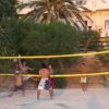 Persone giocano a beach volley su una spiaggia, circondate da vegetazione e palme.