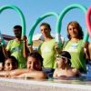 Bambini e adulti in piscina tengono cerchi colorati sopra la testa e sorridono.