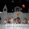Spettacolo teatrale con performer in abiti bianchi e un edificio di scenografia sullo sfondo.