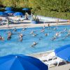 Persone fanno acquagym in piscina con ombrelloni blu.