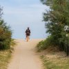 Persona che cammina verso una spiaggia sabbiosa, fiancheggiata da cespugli verdi.