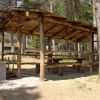 Tavoli da picnic sotto una tettoia di legno in un'area boschiva.
