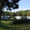 Area campeggio con caravan bianche circondate da alberi e siepi verdi sotto cieli blu.